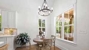 A bright kitchen with white cabinets, a wooden dining table with chairs, a chandelier, and large windows with a view of greenery. - Hickory Cottage - 1bdrm Retreat (Taylors)