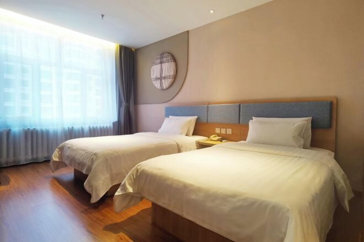 A hotel room with two beds, a large window with curtains, a round wall decoration, and a wooden floor.