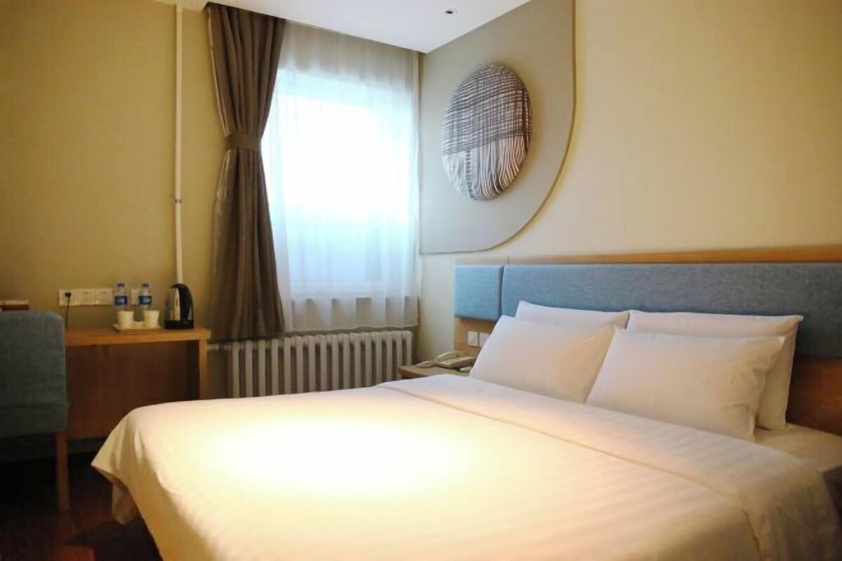 A hotel room with a large bed, a bedside table with a lamp, a window with curtains, and a wall-mounted artwork.