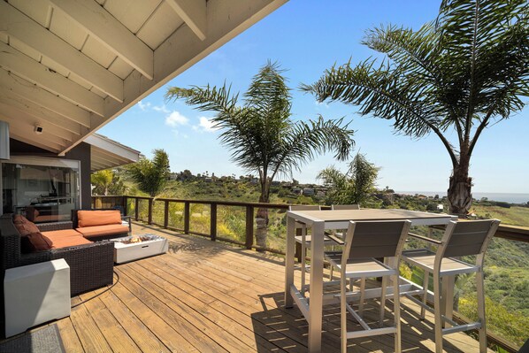 Une terrasse en bois, aménagée avec du mobilier d’extérieur, notamment un canapé et des fauteuils, qui offre une vue imprenable sur un paysage pittoresque, avec des arbres et la mer à l’horizon.