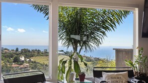 Een kamer met uitzicht op de oceaan, een grote plant en twee stoelen met een tafel ertussen.