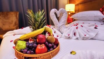 A neatly made bed with a fruit basket, a heart-shaped towel, and rose petals.