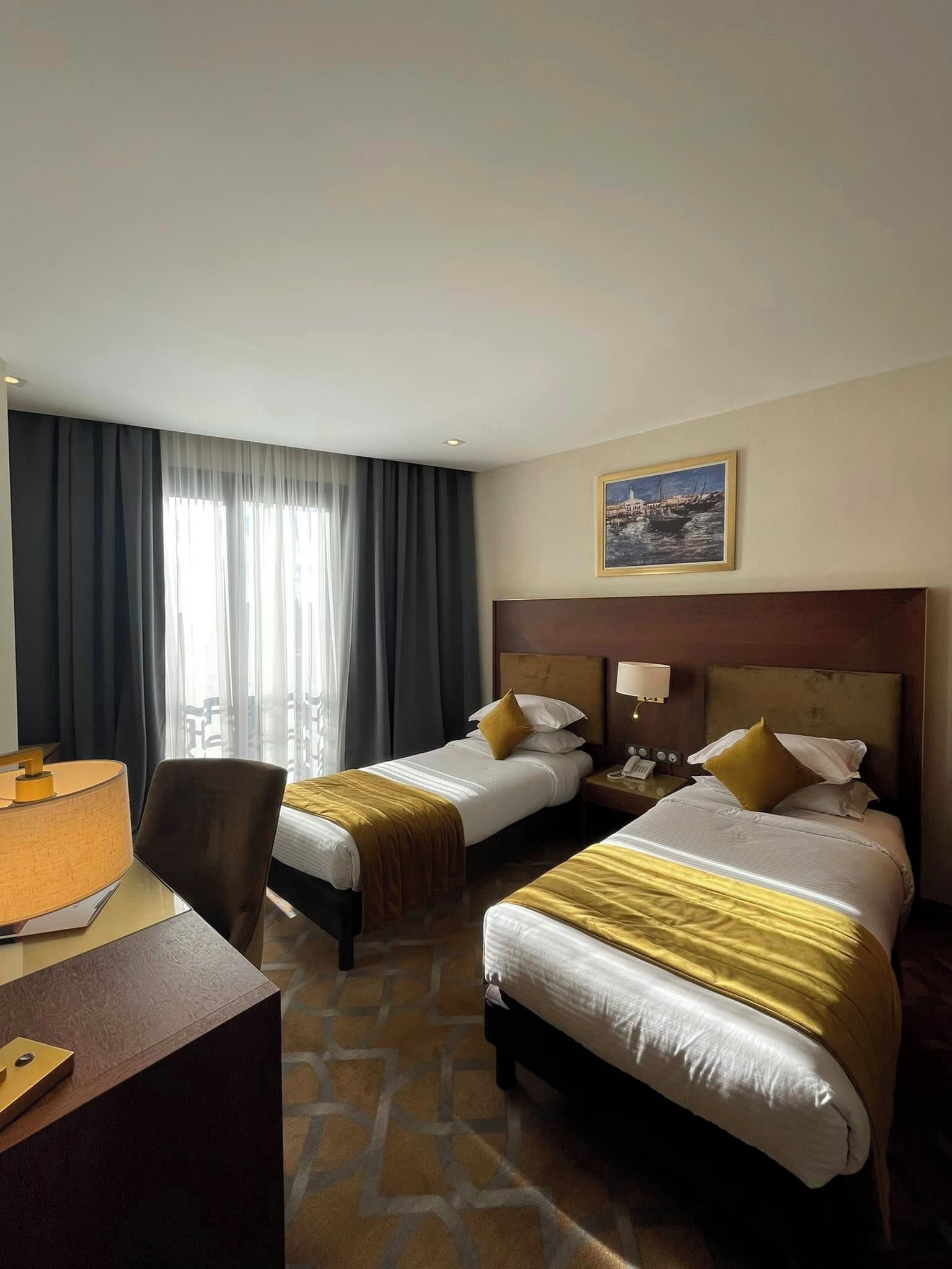 A hotel room with two beds, a desk, and a chair. There is a painting on the wall and a window with curtains.