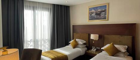 A hotel room with two beds, a desk, and a chair. There is a painting on the wall and a window with curtains.