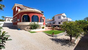 Una casa de dos plantas con fachada roja y nivel superior blanco, un pequeño jardín con césped y plantas en macetas, y un cielo azul despejado.