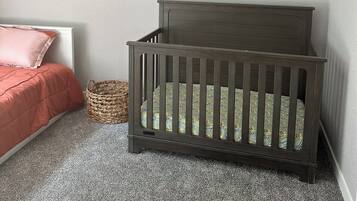A room with a crib, a bed with a pink blanket, a woven basket, and a desert-themed wall art.