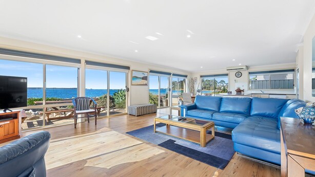 A spacious living room with a large blue sofa, a wooden coffee table, and a television. The room has large windows offering a view of the sea. - Ocean's Edge in Batehaven (Batehaven)