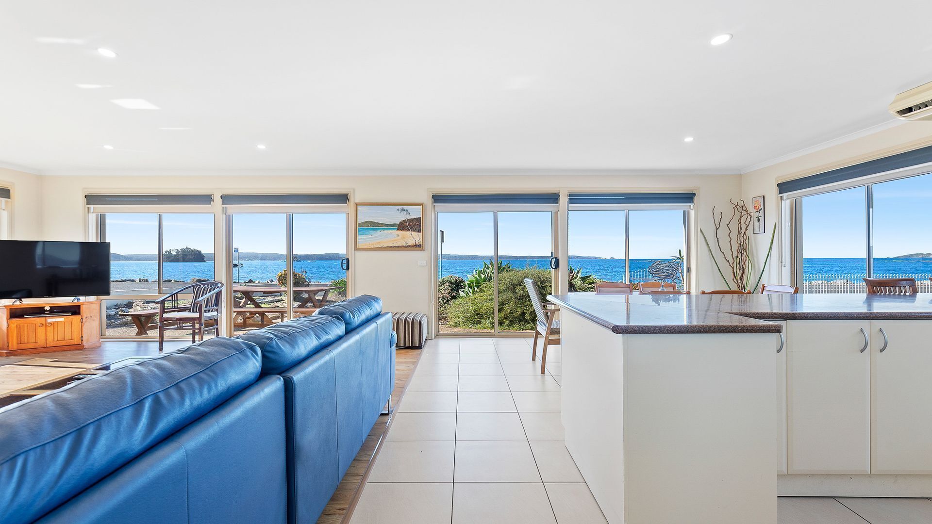 A modern living room with a large TV, blue leather sofa, and a dining area with a view of the sea.