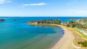 A coastal area with a sandy beach, clear blue waters, and a small island with trees. - Ocean's Edge in Batehaven (Batehaven)
