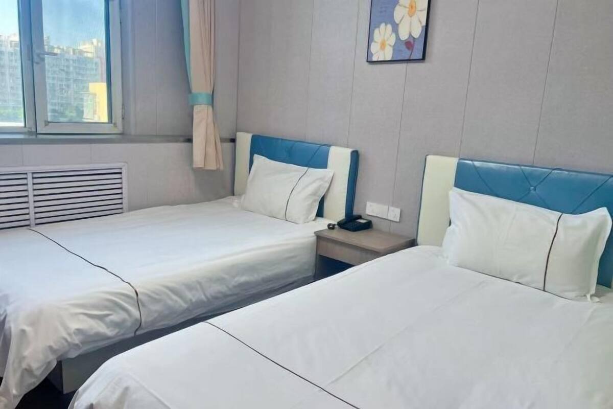 Een hotelkamer met twee bedden, een raam met uitzicht op gebouwen, een kunstwerk aan de muur en een nachtkastje met een telefoon.