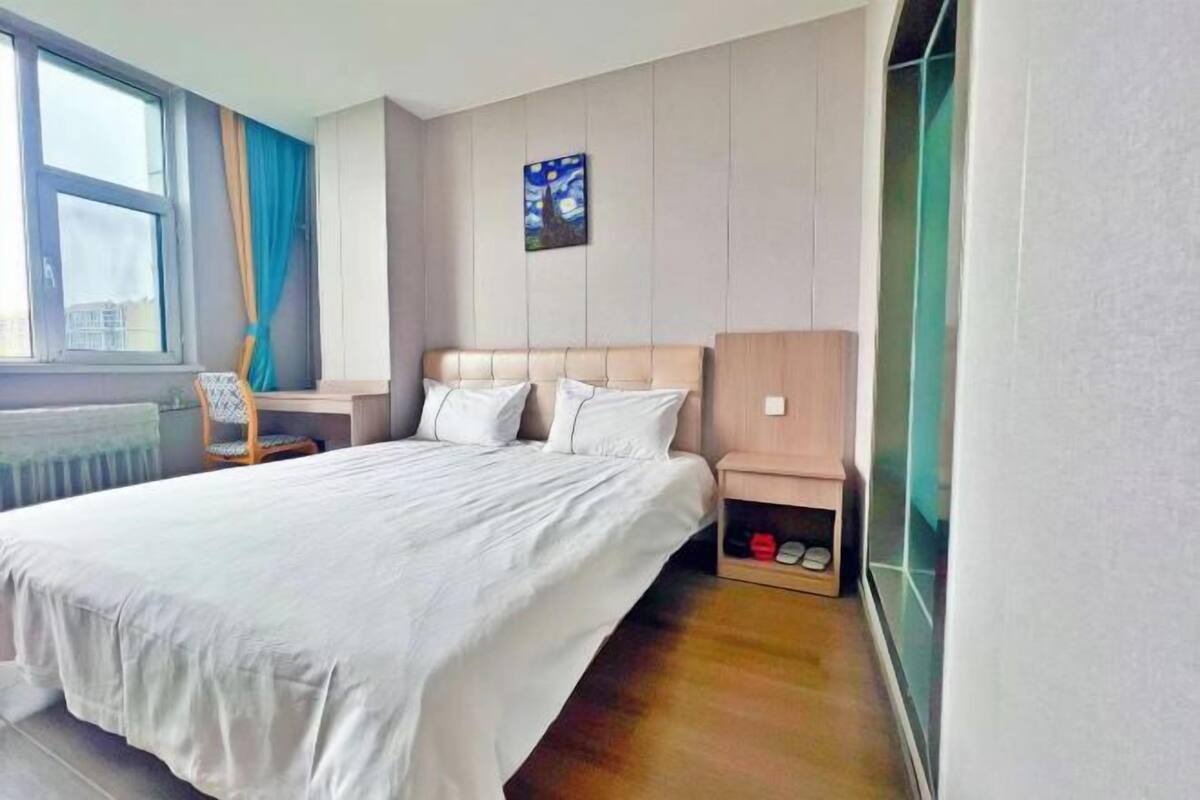 Een hotelkamer met een bed, een nachtkastje, een stoel, een raam met gordijnen, een schilderij aan de muur en een schoenenrek.