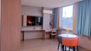 Ett hotellrum med en platt-TV, ett skrivbord, en stol, ett litet bord med en vas och ett stort fönster med gardiner.