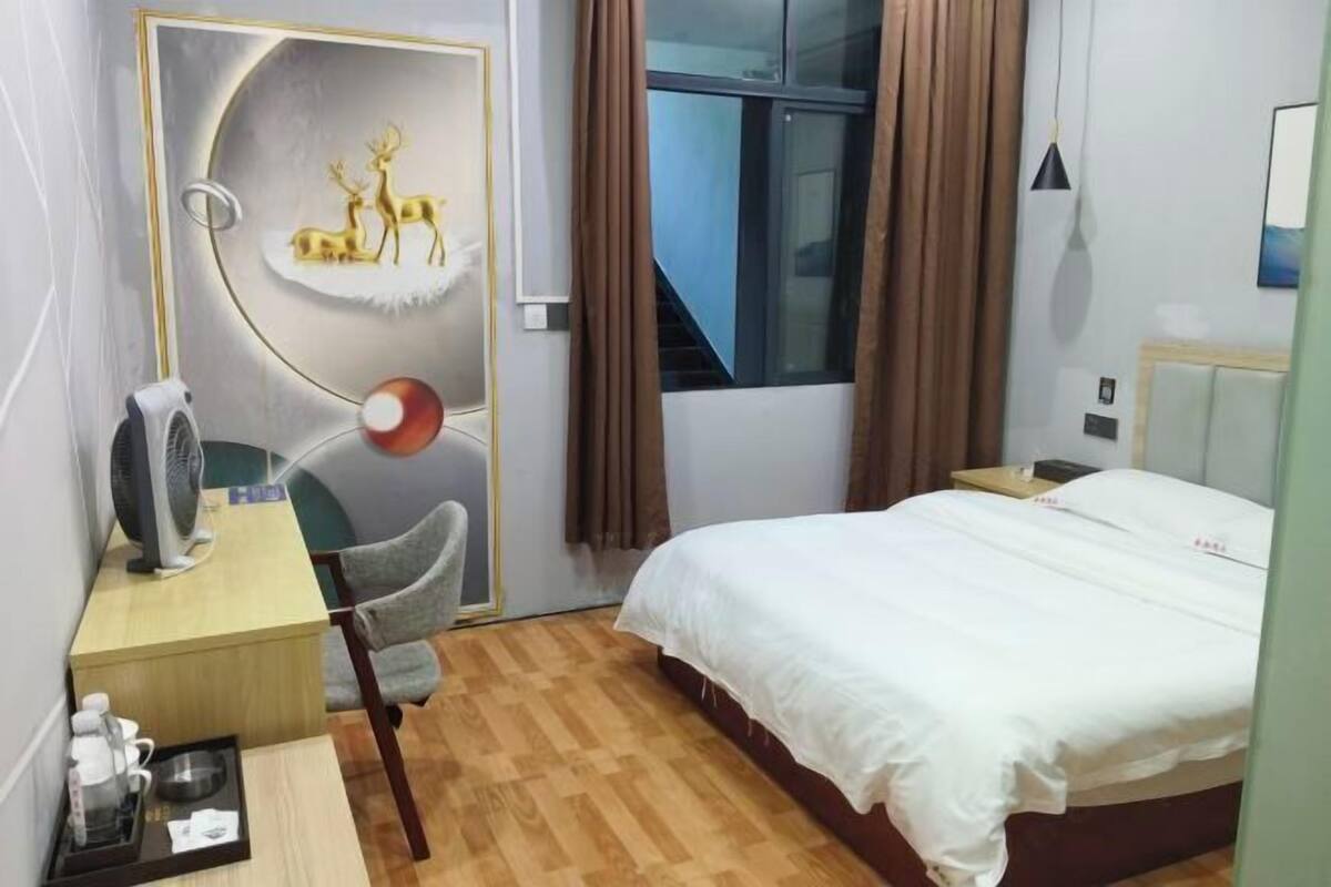 Ein Hotelzimmer mit einem Bett, einem Schreibtisch mit Stuhl, einem Tischventilator, einem Wandfernseher und einem Fenster mit Vorhängen.