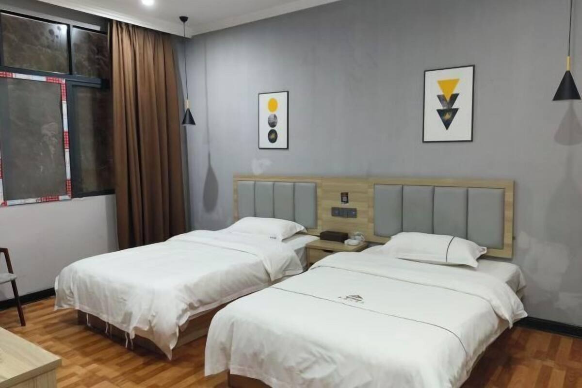 Ein Hotelzimmer mit zwei Betten, einem hölzernen Kopfteil, einem Fenster mit Vorhängen und zwei gerahmten Kunstwerken an der Wand.