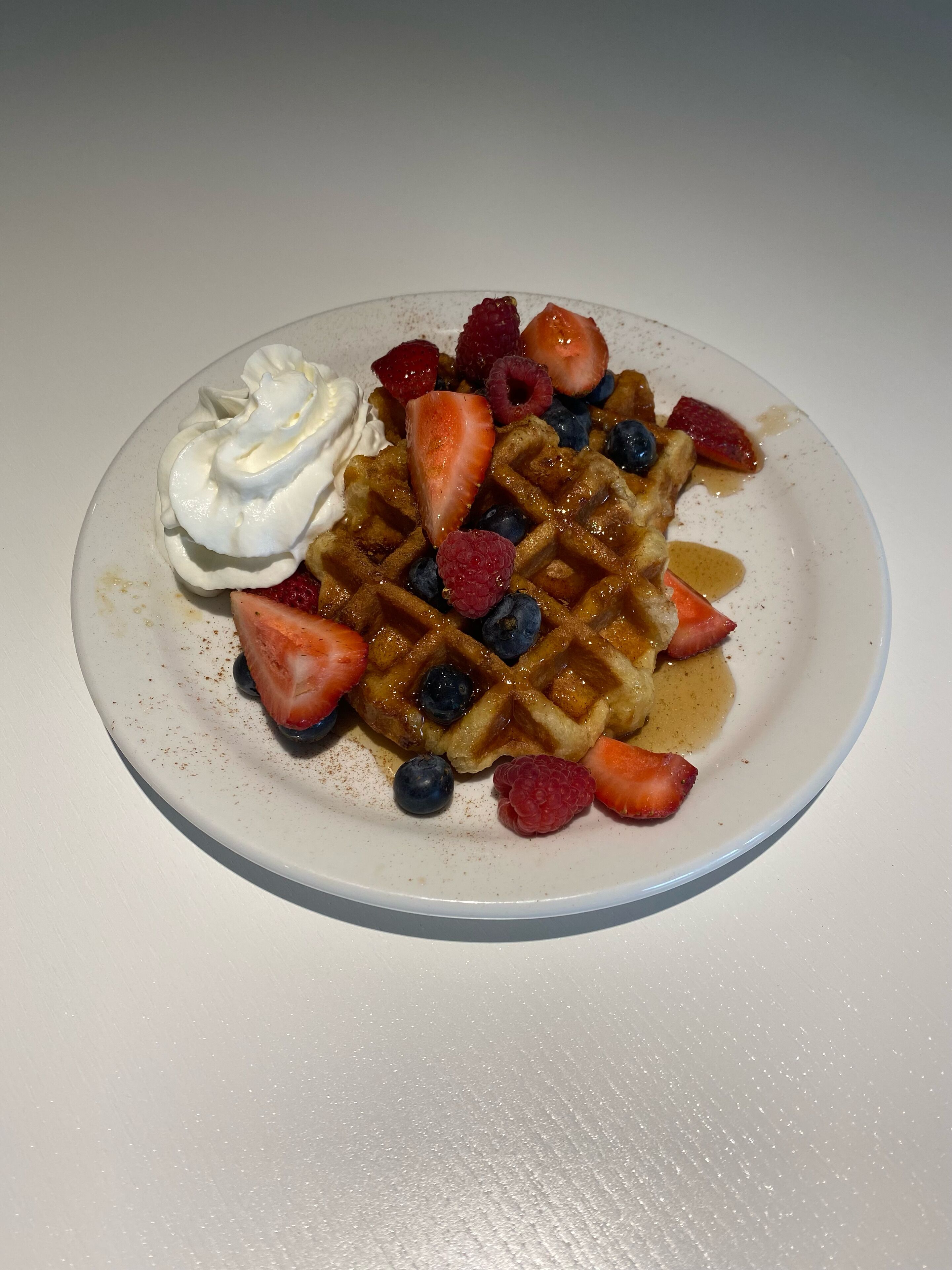A plate with waffles topped with whipped cream, strawberries, blueberries, and raspberries, with syrup on the side.