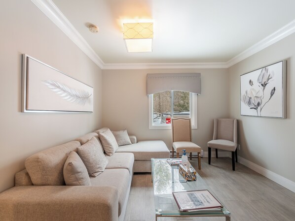 Una sala de estar moderna con un sofá beige, una mesa de centro de vidrio y una ventana con vistas a árboles.