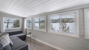 Ein gemütliches Zimmer mit einem grauen Sofa, einem kleinen Tisch mit einer Pflanze und großen Fenstern mit Blick auf eine verschneite Landschaft.