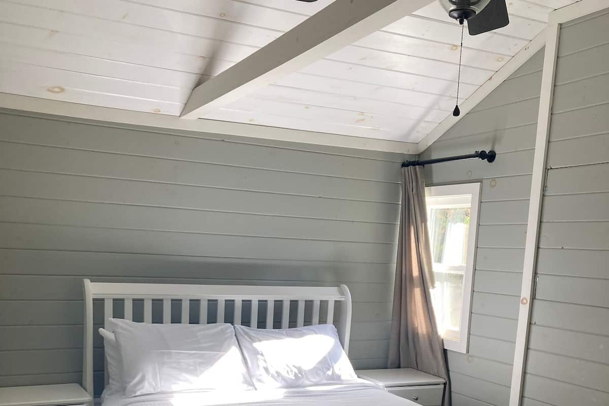 A bedroom with a white bed, a nightstand, a window with curtains, and a ceiling fan.
