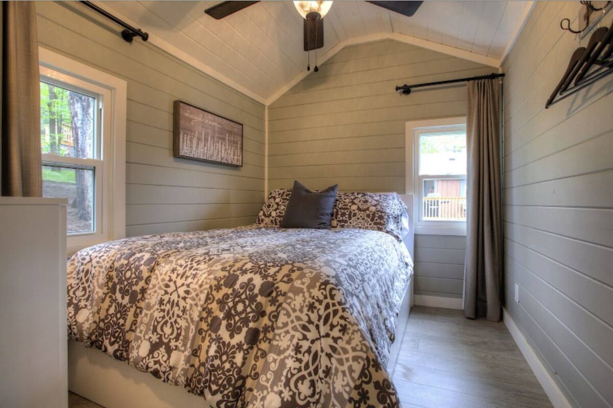 A bedroom with a bed, a ceiling fan, a window with curtains, and a wall-mounted light fixture.