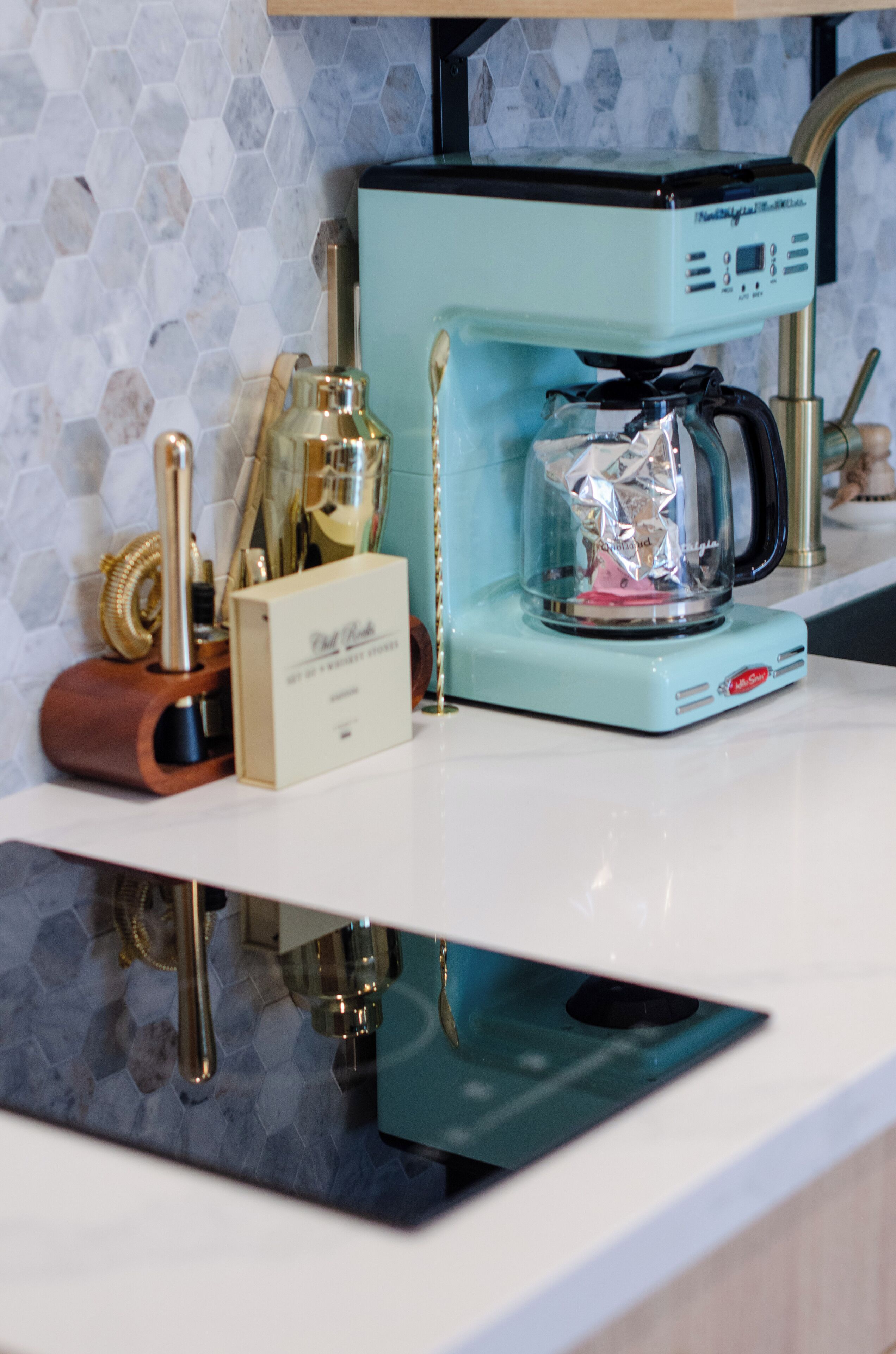 Una cocina con cafetera, un descansaplatos de madera y un recipiente de vidrio sobre una encimera blanca.