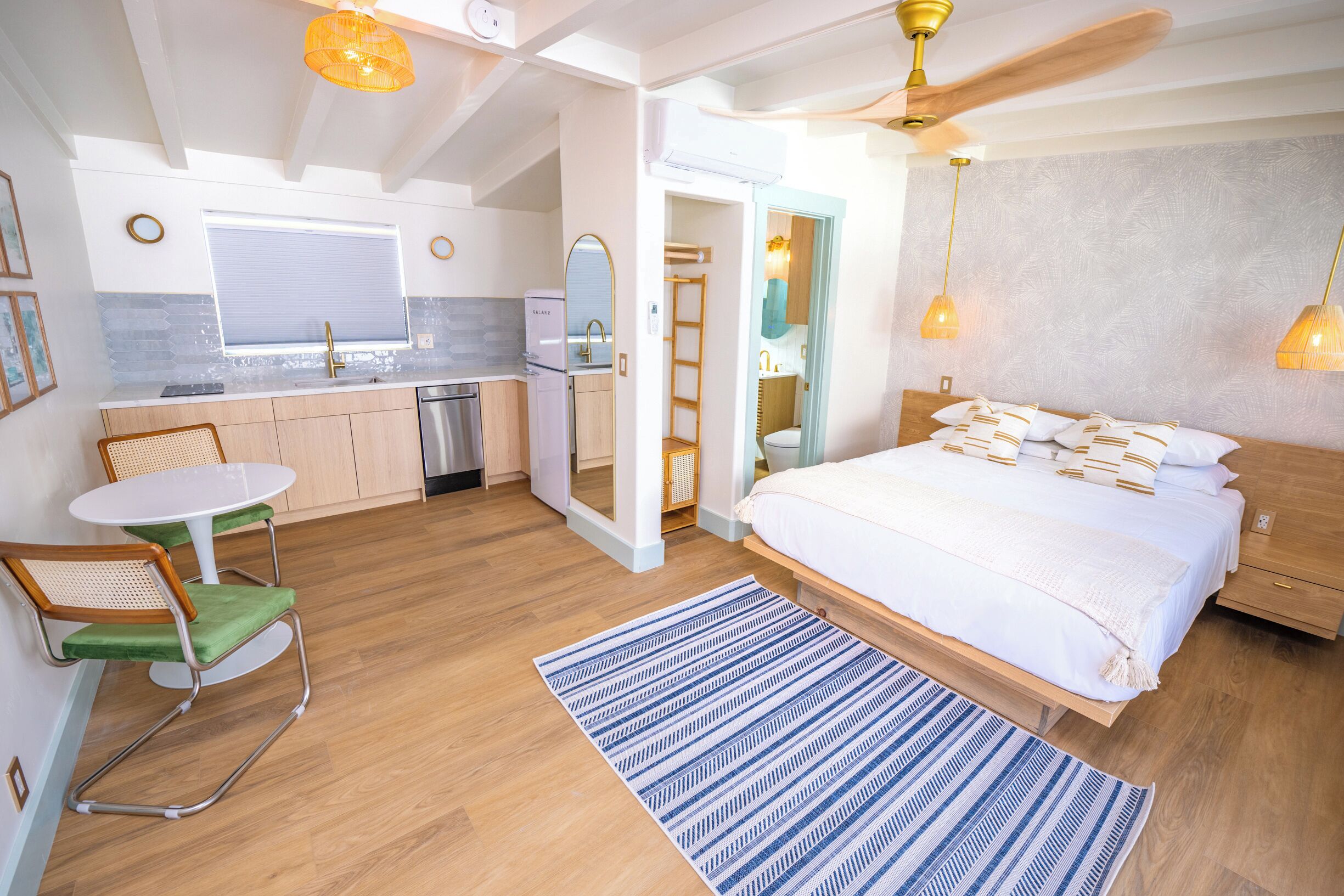 Una habitación de hotel compacta con kitchenette, una cama con ropa blanca, una pequeña mesa redonda con sillas y una alfombra azul a rayas.