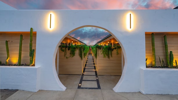 Une entrée architecturale moderne, dotée d’une arche centrale illuminée par des lampes murales, flanquée de jardinières contenant des cactus, et un chemin menant à travers l’arche.