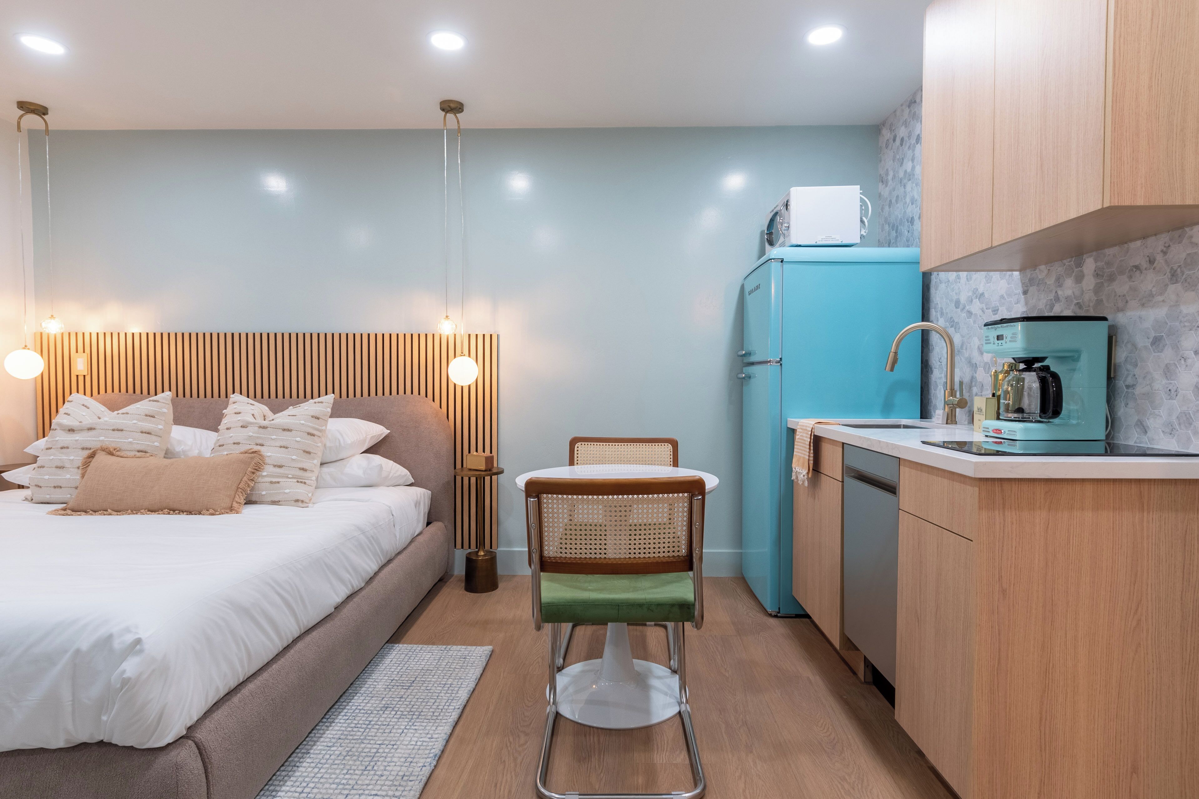 A compact hotel room with a bed, bedside table, chair, and kitchenette.