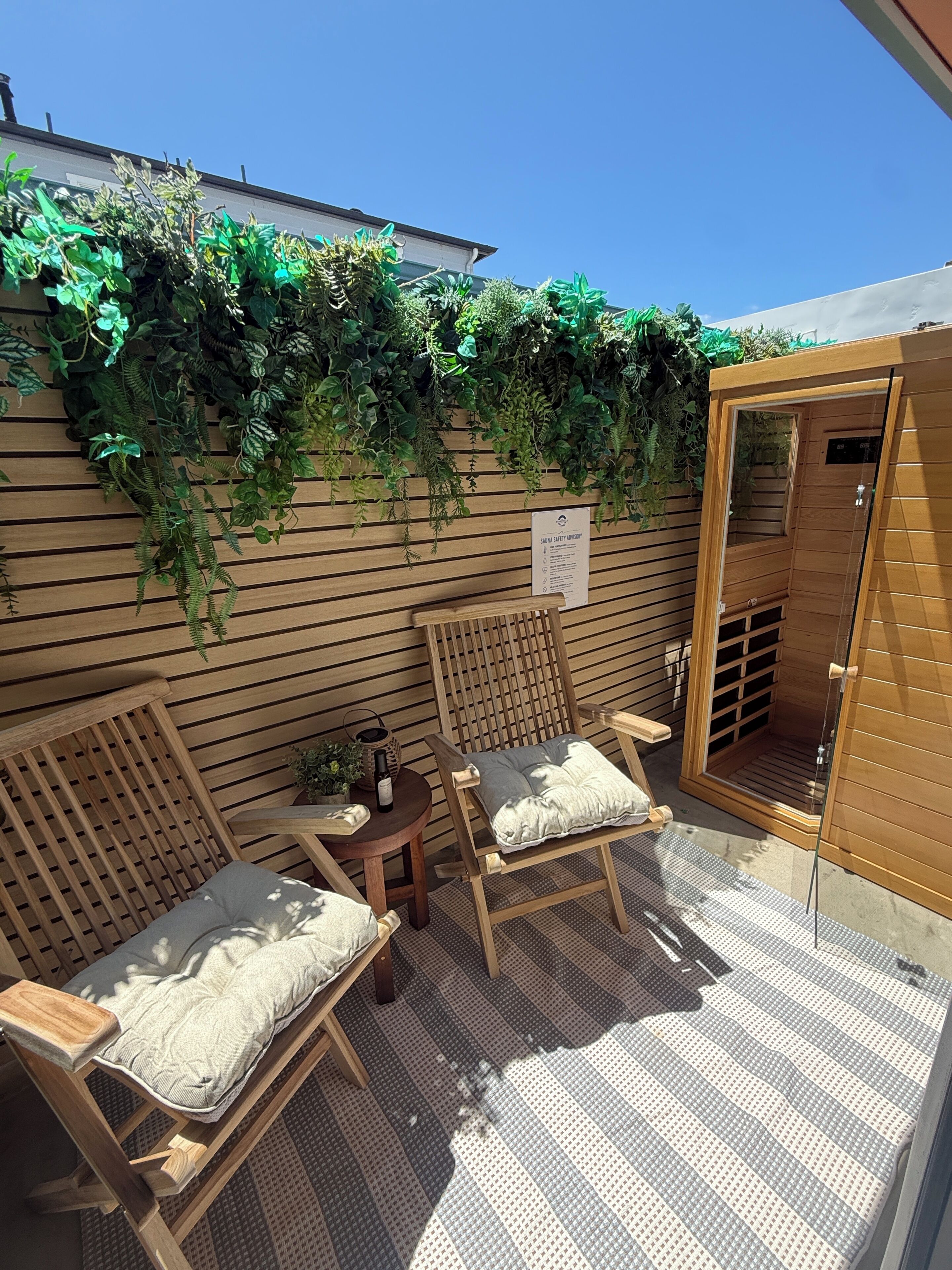 Une terrasse en bois avec deux chaises et une table, une porte de sauna et un mur recouvert de plantes vertes.