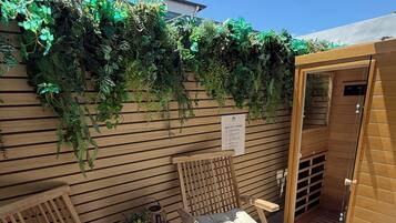 Une terrasse en bois avec deux chaises et une table, une porte de sauna et un mur recouvert de plantes vertes.