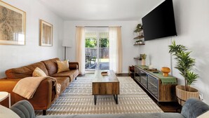 A living room with a brown leather sofa, a wooden coffee table, a flat-screen TV mounted on the wall, and a large window with curtains.