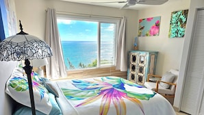 A bedroom with a large bed, a ceiling fan, a view of the sea, and a cabinet with artwork.