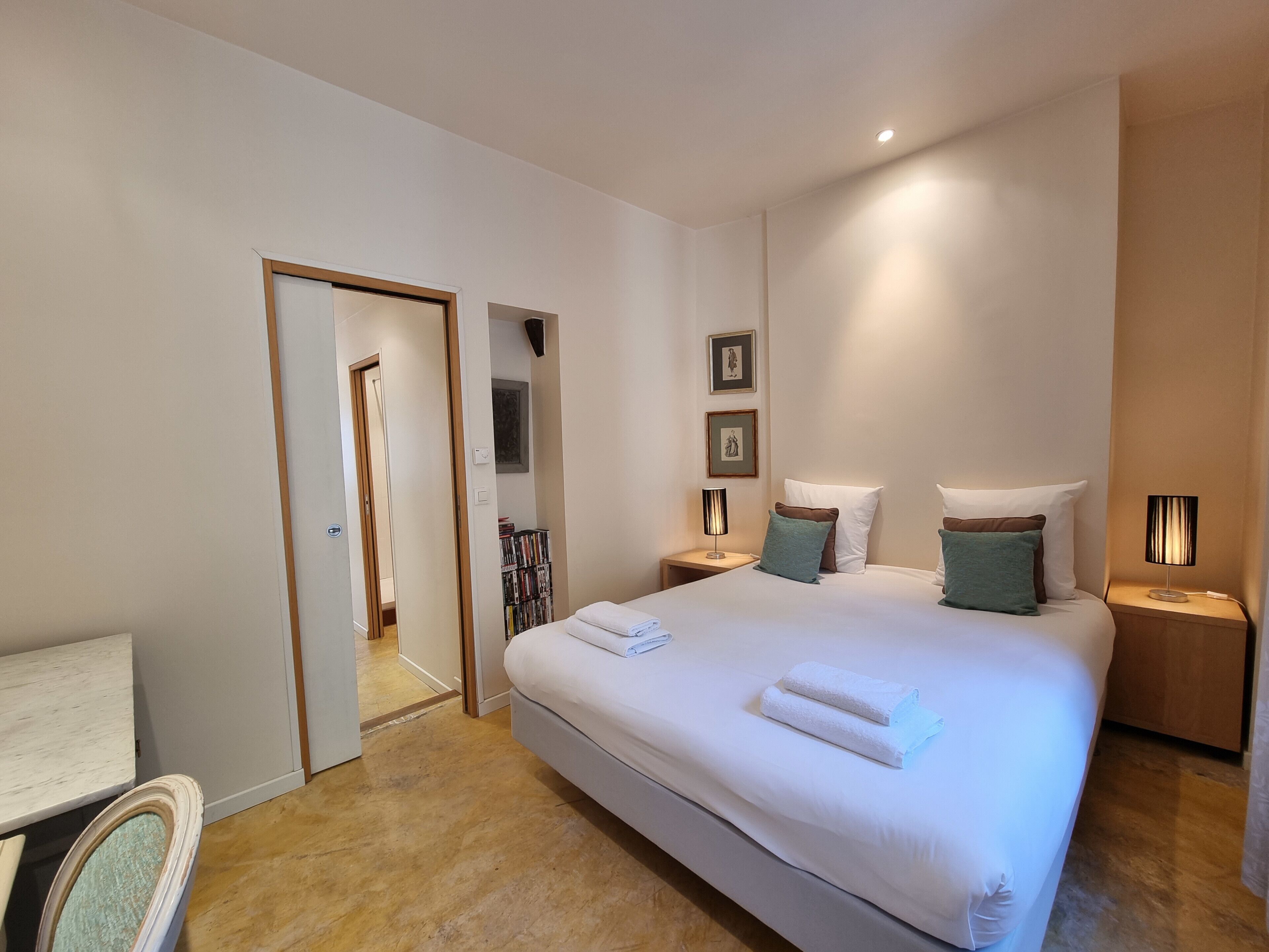 A modern hotel room with a large bed, bedside tables, a bookshelf, and a door leading to another room.