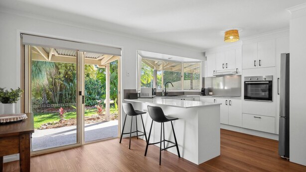 A modern kitchen with white cabinets, a central island with bar stools, and a large window overlooking a garden. - Pet Friendly Marcoola Surfside Escape (Marcoola)