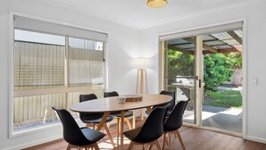 A dining area with a round table, six black chairs, a floor lamp, and a sliding glass door leading to an outdoor space with a grill. - Pet Friendly Marcoola Surfside Escape (Marcoola)