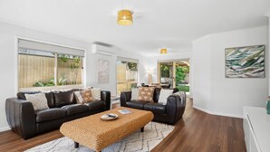 A spacious living room with a leather sofa set, a woven coffee table, and large windows offering views of a backyard. - Pet Friendly Marcoola Surfside Escape (Marcoola)