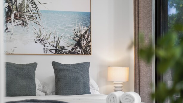 Ein ordentlich bezogenes Bett mit Kissen, einem zusammengerollten Handtuch und einer Wandlampe.