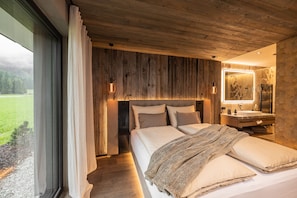 Quarto com cama grande, cabeceira de madeira, banheiro com espelho e vista para uma paisagem verde.