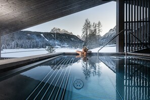 Un jacuzzi con paredes de vidrio y vistas a montañas nevadas, junto a una terraza de madera.