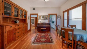 A spacious room with wooden flooring, a piano, a dining table with chairs, and a large window.