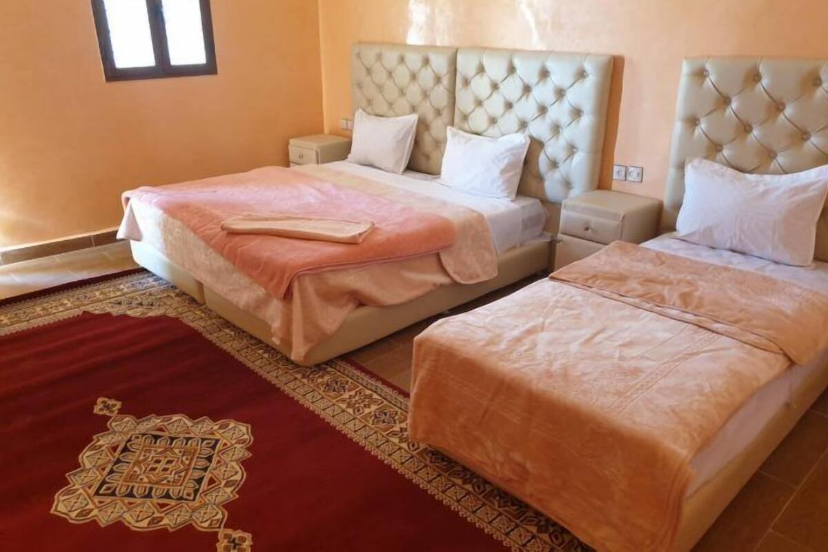 A hotel room with two beds, each with a tufted headboard, a red patterned rug, and a small bedside table.