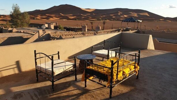 A rooftop terrace with two metal beds and a small round table, set against a desert landscape with sand dunes and a clear sky. - Sahara Garden (Rissani)