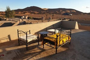 A rooftop terrace with two metal beds and a small round table, set against a desert landscape with sand dunes and a clear sky. - Sahara Garden (Rissani)