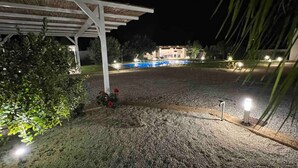 Vista nocturna de un área exterior cubierta con piscina, rodeada de árboles e iluminada por varias luces.