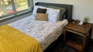 A neatly made bed with floral bedding, a bedside table with a clock, and a window with a view of a building and parked cars. - 2 Bedroom Apartment - Downtown (Marshall)