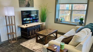 A living room with a sofa, coffee table, TV, and a plant. - 2 Bedroom Apartment - Downtown (Marshall)