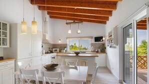 Une cuisine moderne avec des meubles blancs, une table à manger avec des chaises, des luminaires suspendus et une vue à travers une porte-fenêtre coulissante.