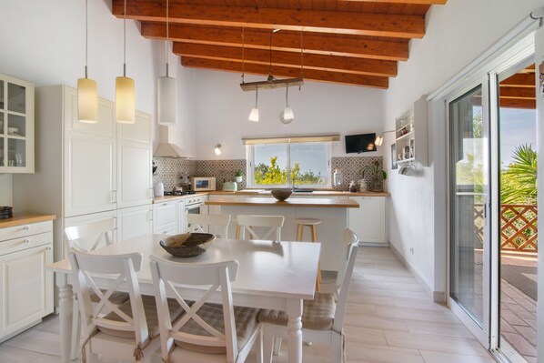 A modern kitchen with white cabinetry, a dining table with chairs, pendant lights, and a view through a sliding glass door.