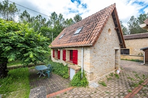 Une maison en pierre avec un toit en tuiles, des volets rouges et un petit jardin agrémenté d’une table et de chaises.