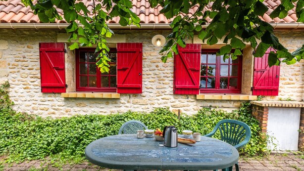 A stone building with red shutters, a round table with chairs, and a small garden area. - Holiday Home “Panissal – Family 4 Guests” with Private Terrace (MONTIGNAC)