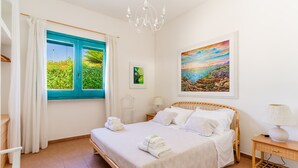 A bedroom with a bed, a nightstand, a lamp, a window with curtains, and a painting on the wall.